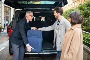  A masked airport transfer chauffeur in a formal suit opens the door of a luxury black sedan for a masked passenger, ensuring a safe and comfortable ride.