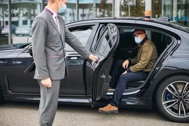 A chauffeur in a suit helps a male traveler load a blue suitcase into the trunk of a black luxury vehicle, while a woman stands nearby.